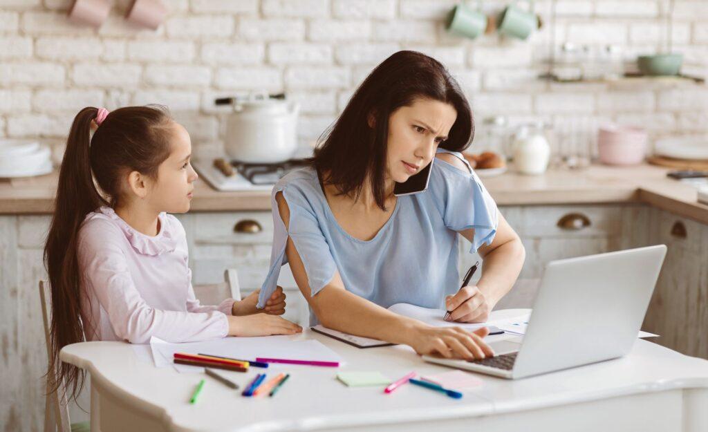 Eine Mutter arbeitet an ihrem Laptop, während sie telefoniert. Ihre kleine Tochter sitzt am Küchentisch und malt mit farbigen Markern. Die Mutter ist eindeutig beschäftigt und versucht sich auf ihre Arbeit zu konzentrieren.