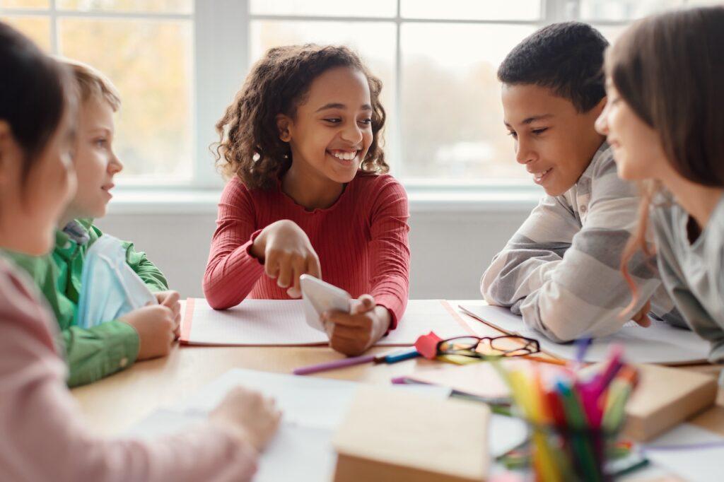 Gruppe von Grundschülern sitzt zusammen an einem Tisch, arbeitet fröhlich an einem Projekt und teilt Ideen im multikulturellen Klassenzimmer.