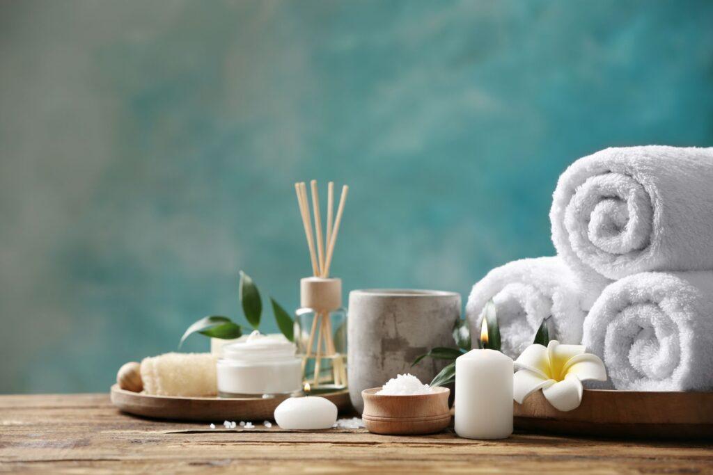 Wellness Accessoires mit Kerzen, weissen Handtuechern und Spa-Duftstaebchen auf Holztisch vor blauem Hintergrund